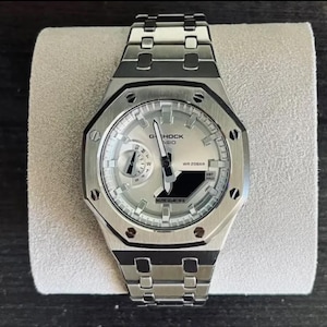 May include: A silver-toned G-Shock Casio wristwatch featuring an octagonal bezel with eight visible screws, a metallic link bracelet, and a light-coloured dial with analogue hands, a day-of-the-week sub-dial, and a digital display window labelled WR 20BAR.