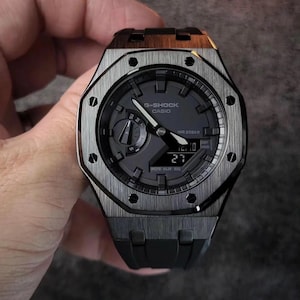 May include: A G-Shock Casio watch with a dark grey face and a black strap. The watch features a brushed metal bezel and a digital display showing the date. Water resistant to 200m.