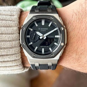 May include: A G-Shock Casio watch with a black and silver octagonal case and a black strap. The watch face is dark grey with white hands and markers, displaying the time as 10:10. Water resistant to 20 bar.