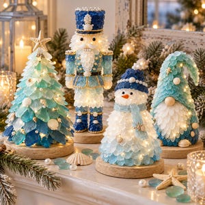 Puede incluir: Un conjunto de cuatro figuras decorativas navideñas sobre bases redondas de madera, que incluye un árbol de Navidad de vidrio marino con una estrella de mar, un cascanueces, un muñeco de nieve y un gnomo, iluminados con luces y con conchas.