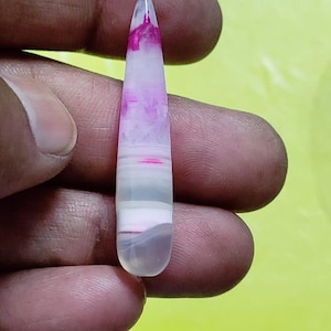 May include: A polished, elongated gemstone with a gradient of colors. The top is a deep magenta, fading to white and gray at the base. The stone is held in a hand, showcasing its smooth, polished surface.