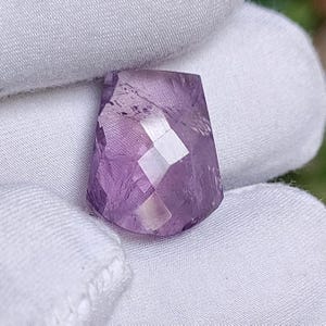 May include: A faceted, trapezoid-shaped amethyst gemstone. The gem is a translucent purple colour with multiple facets that reflect light. The stone is held against a white fabric background.
