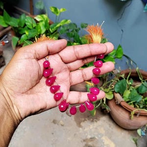 Puede incluir: Un hilo de piedras preciosas translúcidas en forma de lágrima de color rosa fucsia, ensartadas en un alambre fino, con una pequeña borla naranja deshilachada en cada extremo, formando un componente decorativo para joyería.