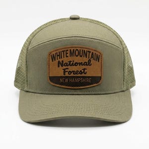 Könnte beinhalten: Olivgrüne Trucker-Mütze mit braunem Lederaufnäher. Der Aufnäher trägt den Text "WHITE MOUNTAIN National Forest NEW HAMPSHIRE". Die Mütze hat einen Mesh-Rücken und einen gebogenen Schirm. Ein klassisches Outdoor-Accessoire.