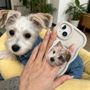 Può includere: Una custodia per smartphone beige con bordo ondulato e un supporto circolare con un ritratto realistico di un piccolo cane bianco e marrone dal pelo arruffato. La custodia presenta un ritaglio per la doppia fotocamera e una finitura opaca.