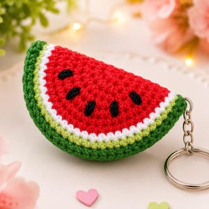 May include: A crochet keyring shaped like a watermelon slice, featuring a red centre with black embroidered seeds, a white border, and a green rind. The item is attached to a silver-toned metal split ring by a short metal chain.