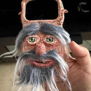 Puede incluir: Un abridor de botellas con un rostro de arcilla texturizado de color marrón rojizo, con ojos verdes, una nariz prominente y una barba y bigote sintéticos de color gris. La parte superior tiene una abertura rectangular para abrir botellas.