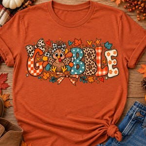 Peut inclure: Un t-shirt à manches courtes orange brûlé avec le mot GOBBLE écrit en grandes lettres à motifs. La lettre O est remplacée par une dinde de dessin animé portant un nœud à carreaux. Le motif inclut des imprimés léopard, carreaux et pois.