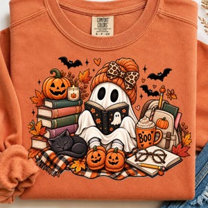 Peut inclure: Un sweat-shirt orange à col rond orné d'un motif représentant un fantôme portant un bonnet et un nœud à imprimé léopard, lisant un livre. Le dessin inclut des livres empilés, une citrouille, un chat noir, une tasse marquée BOO, des lunettes.