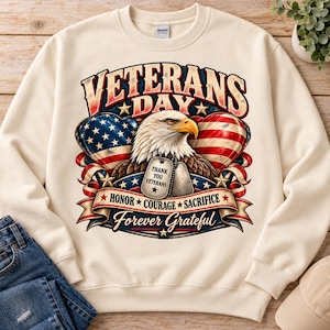 Könnte beinhalten: Ein cremefarbenes Sweatshirt mit Rundhalsausschnitt und einem Grafikdruck eines Weißkopfseeadlers, zwei Erkennungsmarken mit der Aufschrift THANK YOU VETERANS sowie herzförmigen US-Flaggen. Der Text lautet VETERANS DAY, HONOR COURAGE SACRIFICE und Forever Grateful.