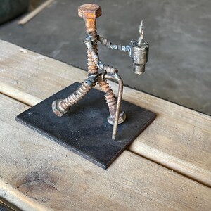 May include: A handcrafted metal sculpture of a person walking, holding a cup. The figure is constructed from various metal hardware pieces, including bolts, screws, and wire, and is mounted on a small, square metal base. The sculpture has a rustic, industrial aesthetic.