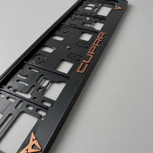 Op de afbeelding: Zwarte kentekenplaathouder met koperkleurige "CUPRA" letters en logo accenten. Het frame heeft een geometrisch ontwerp met diverse uitsparingen en bevestigingspunten. Het koperkleurige logo staat boven en onder het frame.