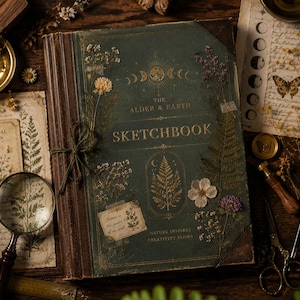 Può includere: Un album da disegno con copertina rigida verde scuro intitolato The Alder & Earth Sketchbook con fasi lunari e illustrazioni botaniche in oro. La copertina è decorata con fiori secchi pressati, un piccolo fiocco di spago e il testo Nature inspires.