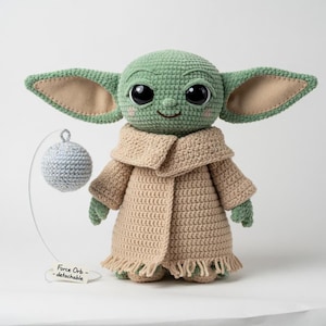 Puede incluir: Mu&ntilde;eco verde tejido a ganchillo con grandes ojos negros, orejas beige y un abrigo beige. Una esfera gris desmontable cuelga de un alambre. El mu&ntilde;eco tiene una expresi&oacute;n sonriente. El texto en el alambre dice "Force Orb - detachable."