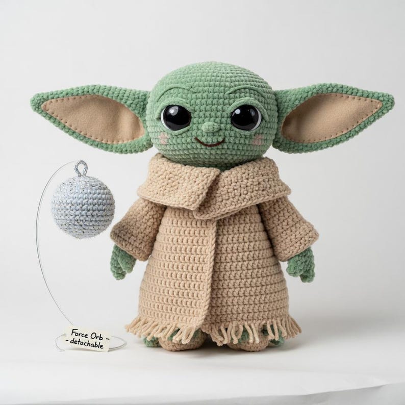 Puede incluir: Mu&ntilde;eco verde tejido a ganchillo con grandes ojos negros, orejas beige y un abrigo beige. Una esfera gris desmontable cuelga de un alambre. El mu&ntilde;eco tiene una expresi&oacute;n sonriente. El texto en el alambre dice "Force Orb - detachable."
