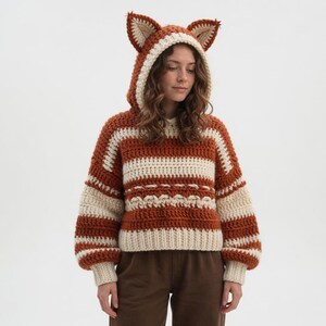 May include: A handmade crochet hoodie in rust and cream colors, featuring a fox ear hood. The sweater has a striped pattern with a decorative stitch detail across the chest. The sleeves are long and the fit is relaxed.
