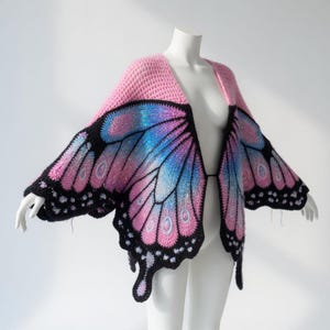 Puede incluir: Cárdigan de ganchillo con alas de mariposa rosa y negro, exhibido en un maniquí. Las alas presentan un degradado de rosa, azul y morado con contornos negros. La parte superior del cárdigan es de color rosa liso.