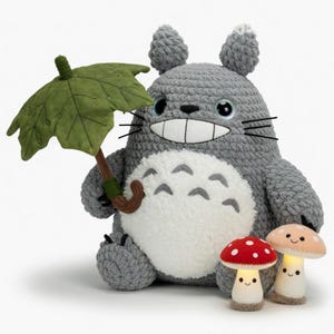 May include: A large, gray crocheted Totoro plushie holding a green leaf umbrella. Two mushroom-shaped night lights with smiling faces are in front of the plushie. One mushroom is red with white spots, and the other is peach.