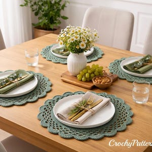 Könnte beinhalten: Ein Holztisch, der für eine Mahlzeit gedeckt ist. Der Tisch ist mit weißen Tellern, grünen Servietten und goldenem Besteck geschmückt, die auf grünen gehäkelten Tischsets liegen. Eine Vase mit Blumen, Trauben und Nüssen vervollständigt das Arrangement.