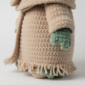 Puede incluir: Mu&ntilde;eca de ganchillo en color beige claro, con un patr&oacute;n de puntada texturizada. La mu&ntilde;eca tiene manos y pies verdes, y una bufanda a juego. La parte inferior de la mu&ntilde;eca tiene un detalle de flecos.