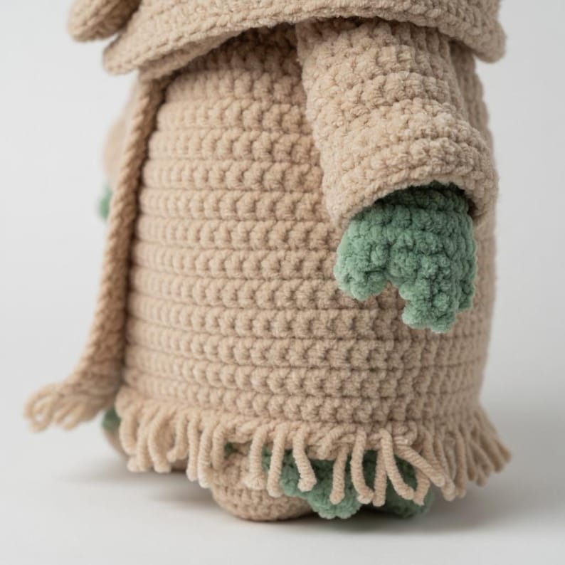 Puede incluir: Mu&ntilde;eca de ganchillo en color beige claro, con un patr&oacute;n de puntada texturizada. La mu&ntilde;eca tiene manos y pies verdes, y una bufanda a juego. La parte inferior de la mu&ntilde;eca tiene un detalle de flecos.