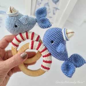 May include: Two crocheted blue whale baby rattles with sailor hats and blue tails. The rattles have wooden rings wrapped in red and white striped yarn. The image is of a handmade baby toy.