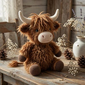 Op de afbeelding: Een handgemaakte bruin en beige gehaakte Highland koeienknuffel met witte hoorns, zittend op een houten oppervlak. De knuffel heeft een pluizig lichaam, een gehaakte snuit en donkere ogen. De achtergrond bevat decoratieve elementen.