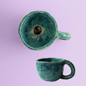 Pode incluir: Duas canecas de cerâmica artesanais com esmalte salpicado turquesa. Uma caneca apresenta um olho esculpido no centro. A outra é uma caneca menor e combinando com uma alça. Ambas as canecas estão em um fundo lavanda.