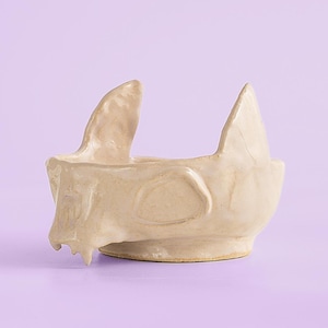 Pode incluir: Uma tigela de cerâmica bege em forma de gato com duas orelhas pontudas e uma abertura arredondada. A tigela tem uma superfície texturizada e assenta numa base redonda. O fundo é de cor lavanda.