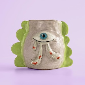 Pode incluir: Um vaso de cerâmica artesanal em forma de rosto estilizado com um único olho azul. O vaso tem uma base cinza com bordas verdes onduladas e detalhes vermelhos. O olho chora lágrimas vermelhas.