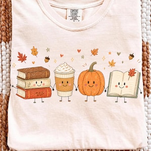 May include: A light peach-colored t-shirt featuring a graphic of a stack of books, a coffee cup, a pumpkin, and an open book, all with smiling faces and thin limbs. The design includes small autumn leaves, acorns, and hearts in a soft color palette.