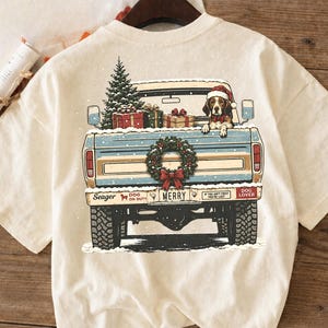 Pode incluir: Uma t-shirt cor de creme com um gráfico da parte traseira de uma carrinha de caixa aberta azul clara a transportar uma árvore de Natal e presentes. Um beagle com um gorro de Pai Natal está sentado na caixa. A porta traseira exibe uma coroa.