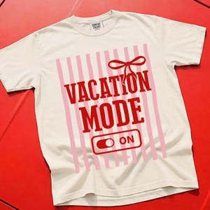 May include: Cream-colored t-shirt with the words "VACATION MODE ON" in red, set against a pink striped background. A red bow is above the text. The shirt is made of a soft material.