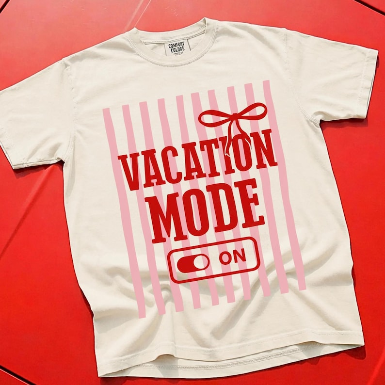May include: Cream-colored t-shirt with the words "VACATION MODE ON" in red, set against a pink striped background. A red bow is above the text. The shirt is made of a soft material.