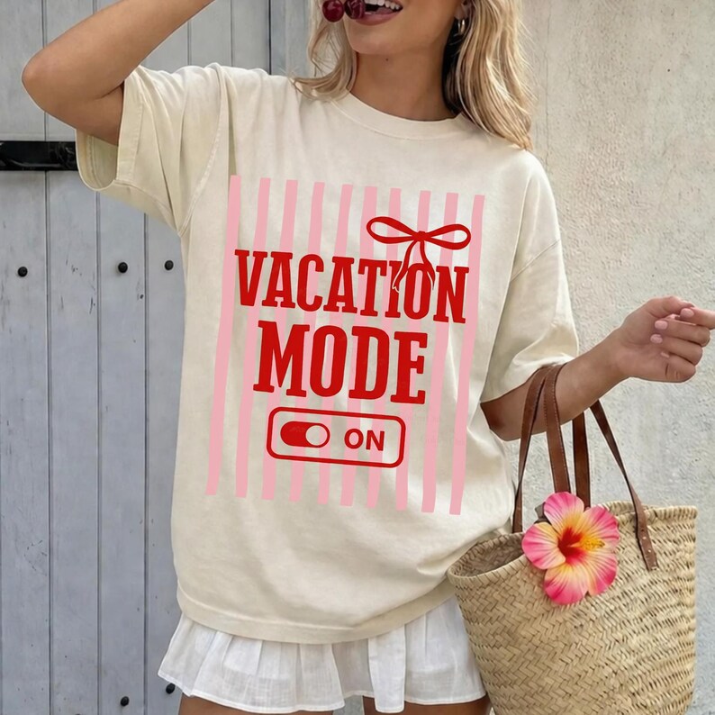 May include: Cream-colored t-shirt with the words "VACATION MODE ON" in red, set against pink vertical stripes. The shirt has a relaxed fit. A straw tote bag with a pink flower is in the image.