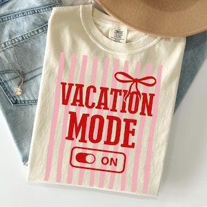 May include: Cream-colored t-shirt with the words "VACATION MODE ON" in red, set against a backdrop of pink vertical stripes. A red bow graphic is above the word "MODE". The shirt is folded, with a tan hat and blue jeans in the background.