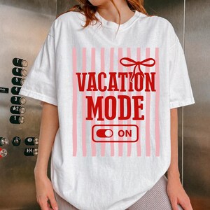 May include: White t-shirt with a red and pink design. The design features the words "VACATION MODE" in bold red letters, with a red bow above and an "ON" switch below. Vertical pink stripes are in the background.
