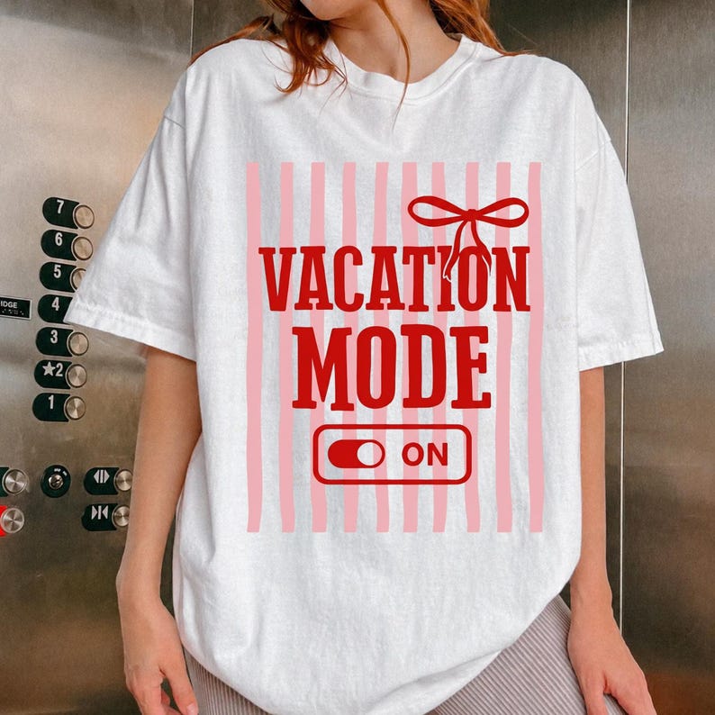 May include: White t-shirt with a red and pink design. The design features the words "VACATION MODE" in bold red letters, with a red bow above and an "ON" switch below. Vertical pink stripes are in the background.