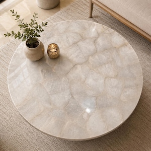 Op de afbeelding: Een ronde salontafel met een oppervlak van gepolijste, doorschijnende witte kwartsstenen tegels in een mozaïekpatroon. Een kleine beige keramische vaas met groen blad en een goudkleurige kaarshouder staan op het tafelblad.