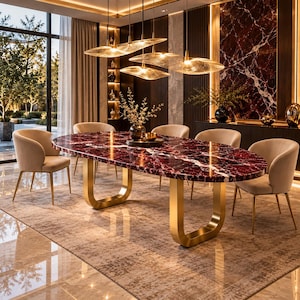 May include: An oval dining table with a deep red marble top and two U-shaped gold metal legs, surrounded by four beige upholstered chairs with gold metal legs. Above the table hang four textured glass pendant lights with gold hardware.