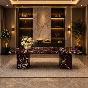 May include: A rectangular dining table featuring a deep red marble top with prominent white veining, supported by two matching block-style marble legs. A white floral arrangement in a metallic vase sits centred on the table surface.
