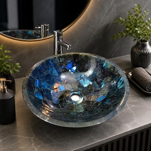Puede incluir: Un lavabo de sobreponer redondo hecho de piedra labradorita pulida, con un mosaico de segmentos en azul profundo, negro y verde azulado iridiscente. El cuenco tiene una superficie lisa y reflectante con un desagüe central y grifo cromado.