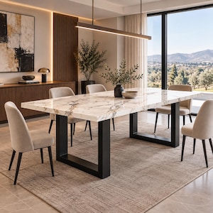 Op de afbeelding: Een rechthoekige eettafel met een wit marmeren blad met grijze en gouden aders, ondersteund door twee dikke zwarte U-vormige metalen poten. De tafel is omringd door vier beige gestoffeerde stoelen met donkere houten poten op een vloerkleed.