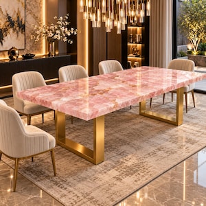 Peut inclure: Une table à manger rectangulaire dotée d'un plateau en pierre de quartz rose poli aux motifs cristallins naturels, soutenue par deux pieds épais en métal doré brossé en forme de U, accompagnée de quatre chaises beiges capitonnées verticalement.