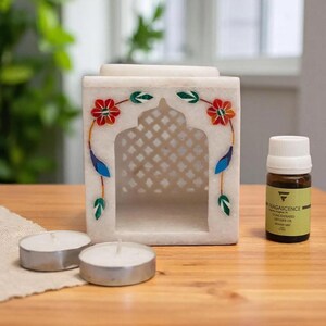 Pode incluir: Um queimador de óleo de mármore branco com um design de treliça recortada e motivos florais incrustados em vermelho, laranja, azul e verde. O conjunto inclui duas velas de chá brancas redondas e um frasco de vidro castanho com o rótulo Fragascence Concentrated Diffuser Oil.