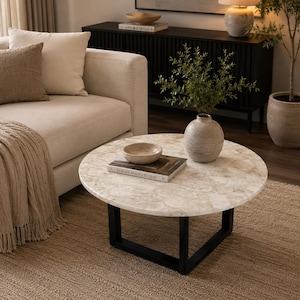 Puede incluir: Una mesa de centro redonda con una superficie de piedra texturizada en color beige claro con un patrón circular y una base de metal negro. Sobre la mesa hay un cuenco beige pequeño y poco profundo y un libro titulado Remote Places to Stay.