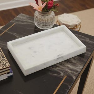 May include: A rectangular white marble tray with subtle grey veining and raised edges. The tray features a smooth, polished surface and a solid construction, providing a clean and neutral base for organizing small items or displaying decorative objects.