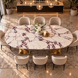 Op de afbeelding: Een ovale eettafel met een wit en paars marmeren blad, ondersteund door twee goudkleurige, X-vormige metalen onderstellen. De tafel is omringd door acht gestoffeerde beige stoelen met gebogen rugleuningen, geplaatst op een groot vloerkleed met patroon.