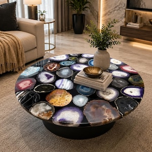 May include: A round coffee table featuring a tabletop composed of numerous multicolored agate slices embedded in a dark resin base. The surface displays various shades of purple, blue, pink, brown, and black, with a small brass bowl and two books.