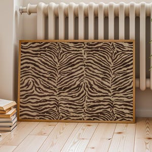 May include: A rectangular wall art piece featuring a dark brown and beige zebra stripe pattern, enclosed within a thin light-colored wooden frame. The artwork is positioned horizontally, displaying a repeating animal print design across its surface.